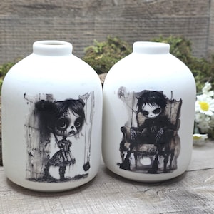 May include: Two white ceramic vases with a matte finish. Each vase features a black and white illustration of a doll-like figure. One vase depicts a girl, the other a boy seated in a chair. The vases are displayed on a wooden surface.