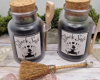 May include: Two glass jars filled with black salt, each with a cork stopper and a label that reads "Black Salt". A small wooden spoon is attached to each jar. A miniature broom with a heart-shaped charm and rose quartz crystals are in front.