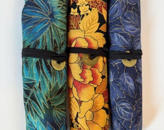 May include: Three fabric pen rolls with unique patterns. One is teal with gold leaf designs, another features large yellow and red flowers, and the third has blue leaves with gold accents. Each roll is secured with a black cord and a decorative coin.