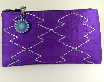 May include: A purple silk zippered pouch with a silver embroidered diamond pattern. The pouch has a silver zipper pull with a blue and green circular charm.