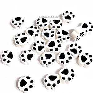 May include: A collection of white paw print beads with black spots scattered on a white background. The beads are made of a smooth material and are designed to resemble a dog's paw. The text "Bead and Craft Stash" is in the upper left corner.
