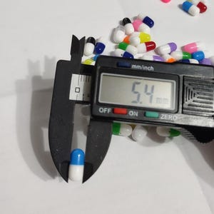 May include: A close-up of a blue and white capsule being measured with a digital caliper. The caliper reads 5.4 mm. The capsule is surrounded by a variety of colorful capsules.