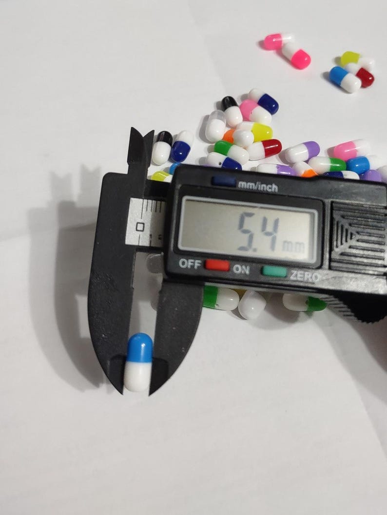 May include: A close-up of a blue and white capsule being measured with a digital caliper. The caliper reads 5.4 mm. The capsule is surrounded by a variety of colorful capsules.