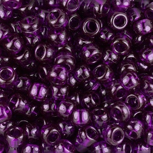 May include: Close-up of a pile of translucent purple pony beads. The beads are cylindrical with a large hole running through the center. The beads have a glossy finish and reflect light, creating highlights and shadows.