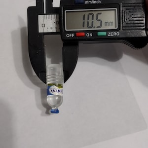 Puede incluir: Una botella de agua en miniatura de pl&aacute;stico transparente con una tapa azul y una etiqueta azul y blanca que dice "Volvic". La botella se est&aacute; midiendo con un calibre digital que lee 10,5 mm.