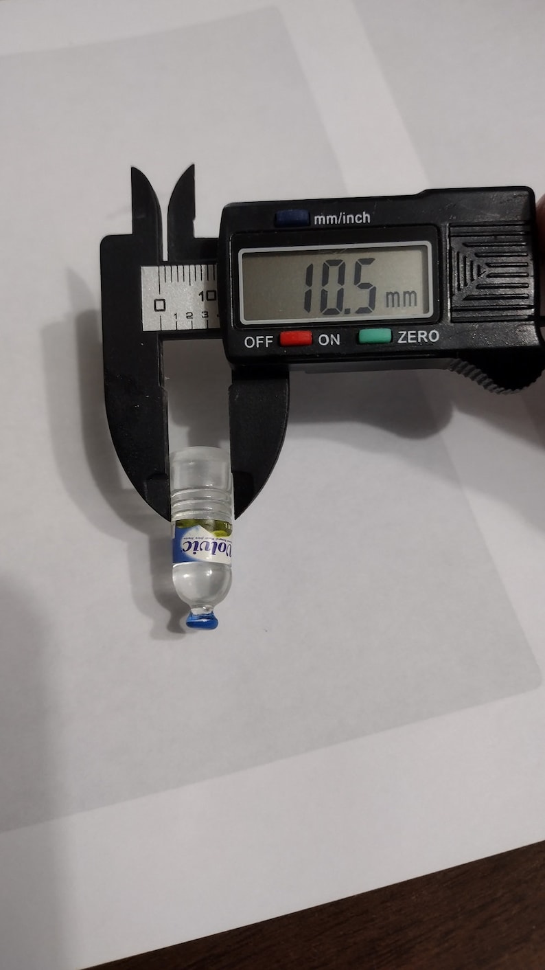 Puede incluir: Una botella de agua en miniatura de pl&aacute;stico transparente con una tapa azul y una etiqueta azul y blanca que dice "Volvic". La botella se est&aacute; midiendo con un calibre digital que lee 10,5 mm.