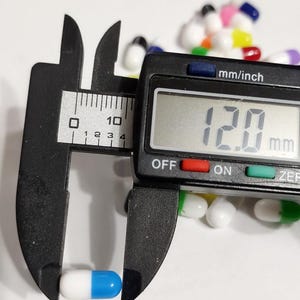 May include: A blue and white capsule pill being measured with a digital caliper. The caliper reads 12.0 mm.