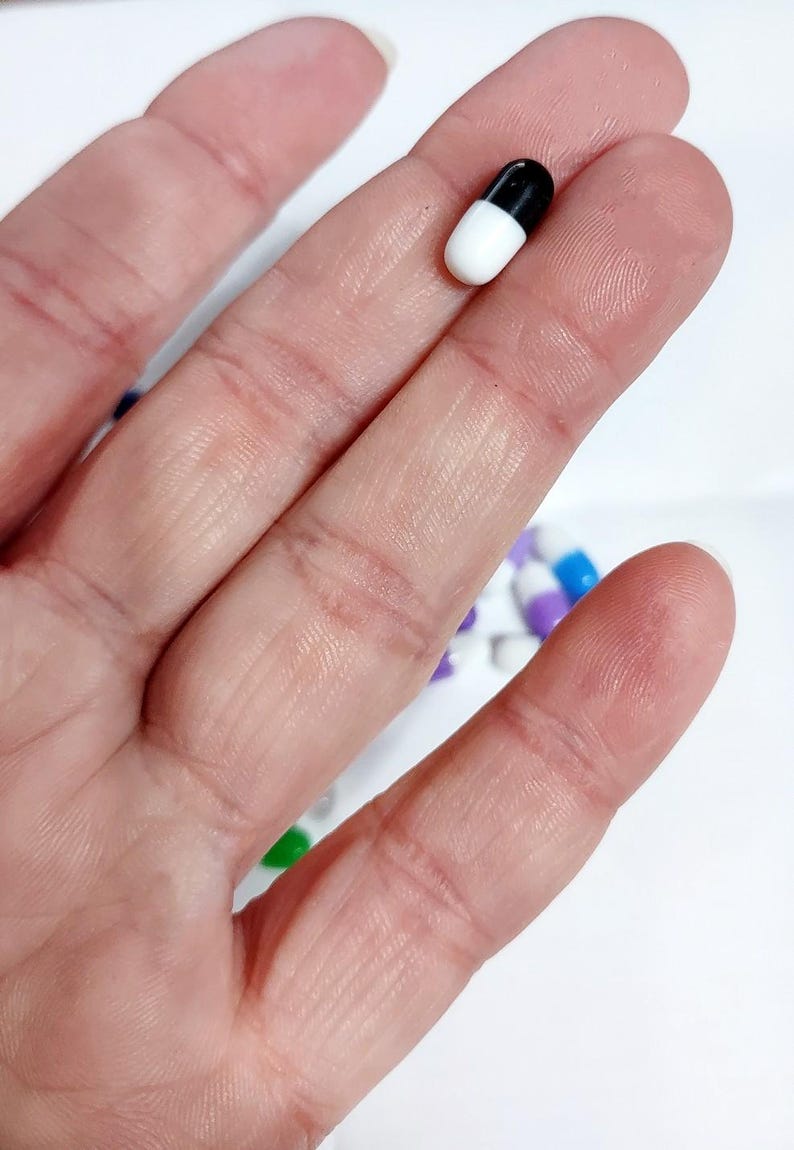 May include: A white and black capsule pill held between two fingers.