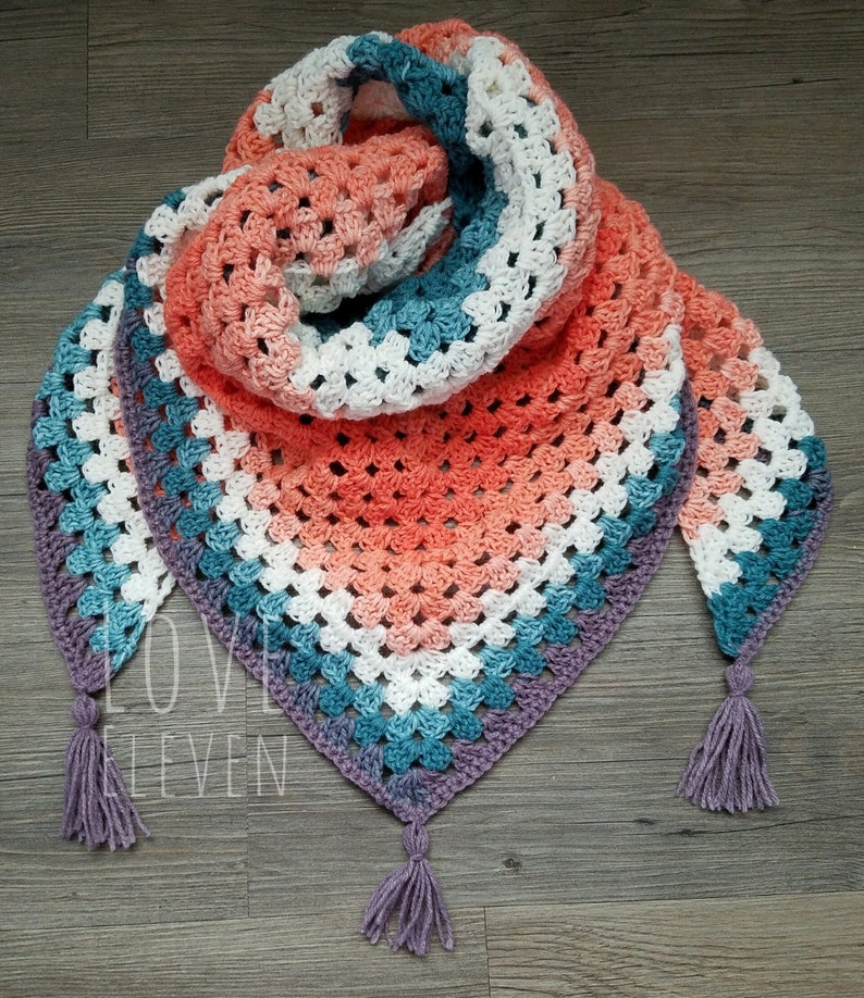 Op de afbeelding: Een driehoekige gehaakte sjaal met een gradi&euml;nt van koraal, wit, turquoise en paars. De sjaal heeft aan elk uiteinde een franje.