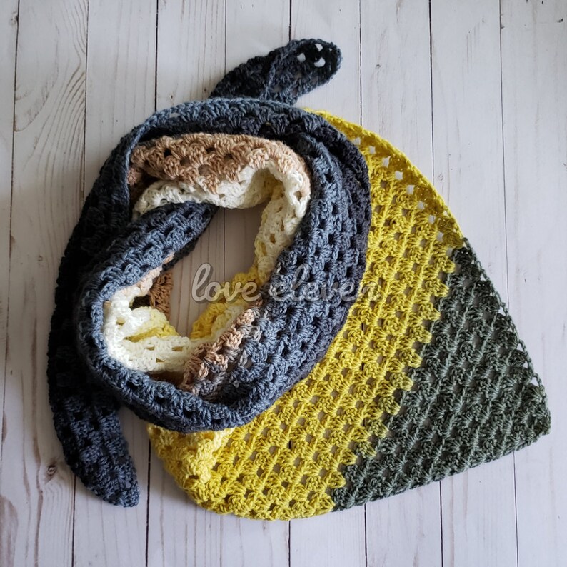 May include: A crocheted triangular scarf in shades of grey, yellow, and green. The scarf is made with a lacy stitch and has a fringed edge.