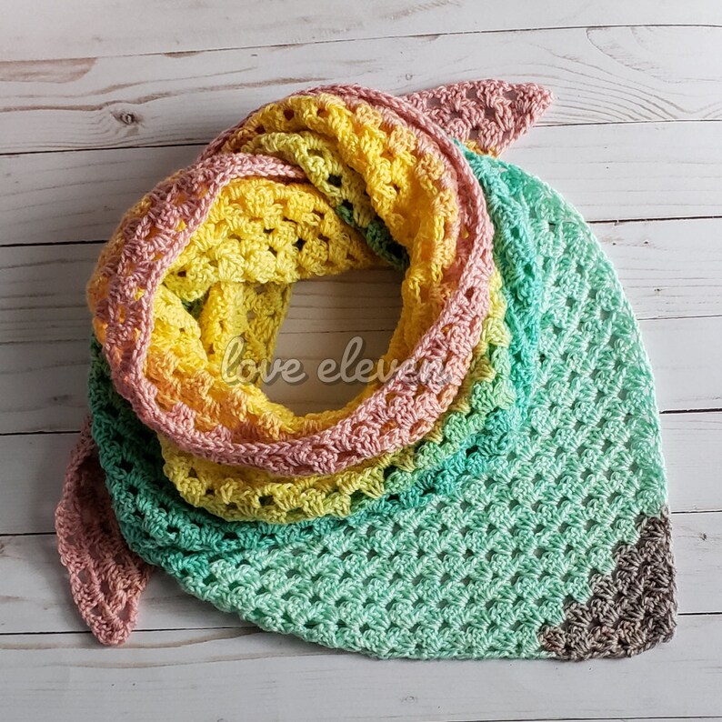 May include: A crocheted triangular shawl in shades of pink, yellow, and green. The shawl is made with a delicate open stitch pattern and has a brown crocheted border at the tip.