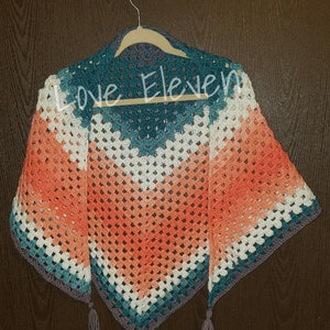 Op de afbeelding: Een gehaakte driehoekige sjaal met een gradi&euml;nt van perzik, wit, turquoise en grijs. De sjaal heeft kwastjes aan de onderste hoeken en hangt aan een houten hanger. De tekst "Love Eleven" is zichtbaar op de sjaal.