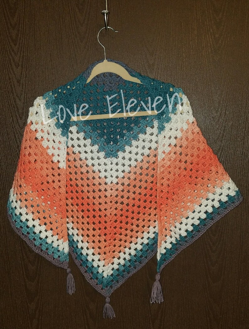 Op de afbeelding: Een gehaakte driehoekige sjaal met een gradi&euml;nt van perzik, wit, turquoise en grijs. De sjaal heeft kwastjes aan de onderste hoeken en hangt aan een houten hanger. De tekst "Love Eleven" is zichtbaar op de sjaal.