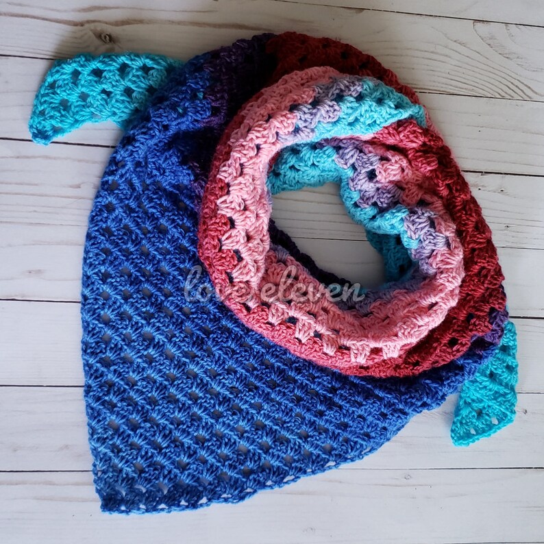 May include: A crocheted triangular shawl with a colourful ombre design. The shawl is made with a variety of shades of blue, pink, purple, and teal yarn. The shawl is folded in half and the edges are visible.