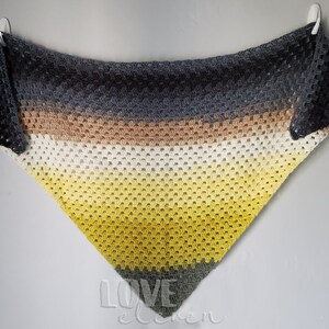 May include: A triangular crocheted shawl with a gradient of colours, starting with dark grey, transitioning to brown, then white, and ending with yellow. The shawl is hanging on a white wall.