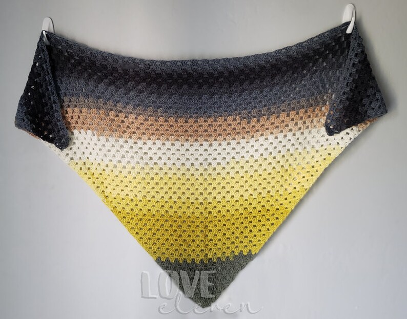 May include: A triangular crocheted shawl with a gradient of colours, starting with dark grey, transitioning to brown, then white, and ending with yellow. The shawl is hanging on a white wall.