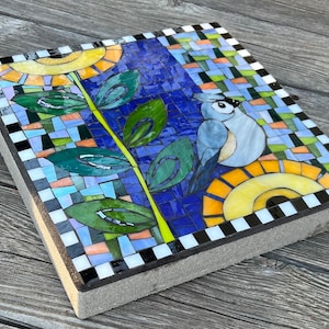 May include: Square mosaic stepping stone featuring a blue bird, sunflowers, and foliage. The design incorporates a blue background with colorful mosaic tiles, green leaves, and a black and white checkered border. The stepping stone is made of concrete.