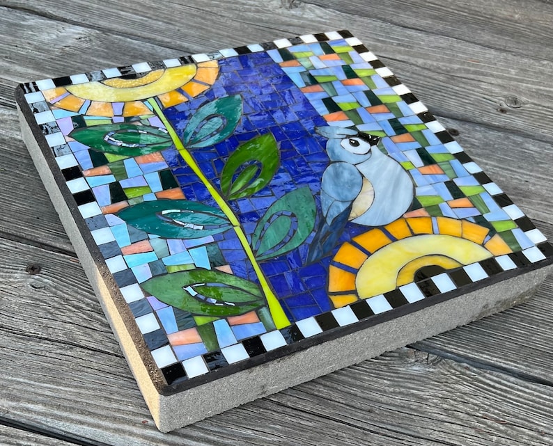May include: Square mosaic stepping stone featuring a blue bird, sunflowers, and foliage. The design incorporates a blue background with colorful mosaic tiles, green leaves, and a black and white checkered border. The stepping stone is made of concrete.
