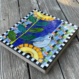 May include: A square mosaic tile featuring a blue bird with a crest, a yellow sun, and green leaves against a blue background. The mosaic is bordered with a black and white checkerboard pattern. The tile is approximately 12 inches square.