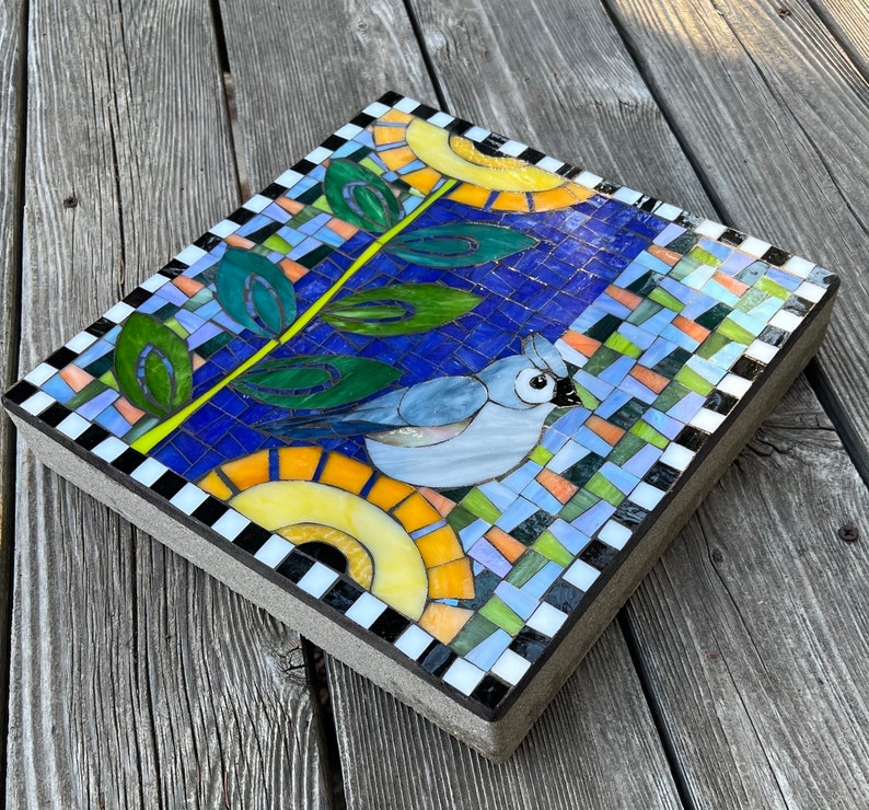 May include: A square mosaic tile featuring a blue bird with a crest, a yellow sun, and green leaves against a blue background. The mosaic is bordered with a black and white checkerboard pattern. The tile is approximately 12 inches square.