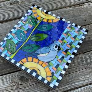 May include: Square mosaic art piece featuring a blue bird, leaves, and sun-like shapes. The mosaic is made of small, colorful tiles, with a blue background. The border is black and white checkered. The artwork is on a wooden base.