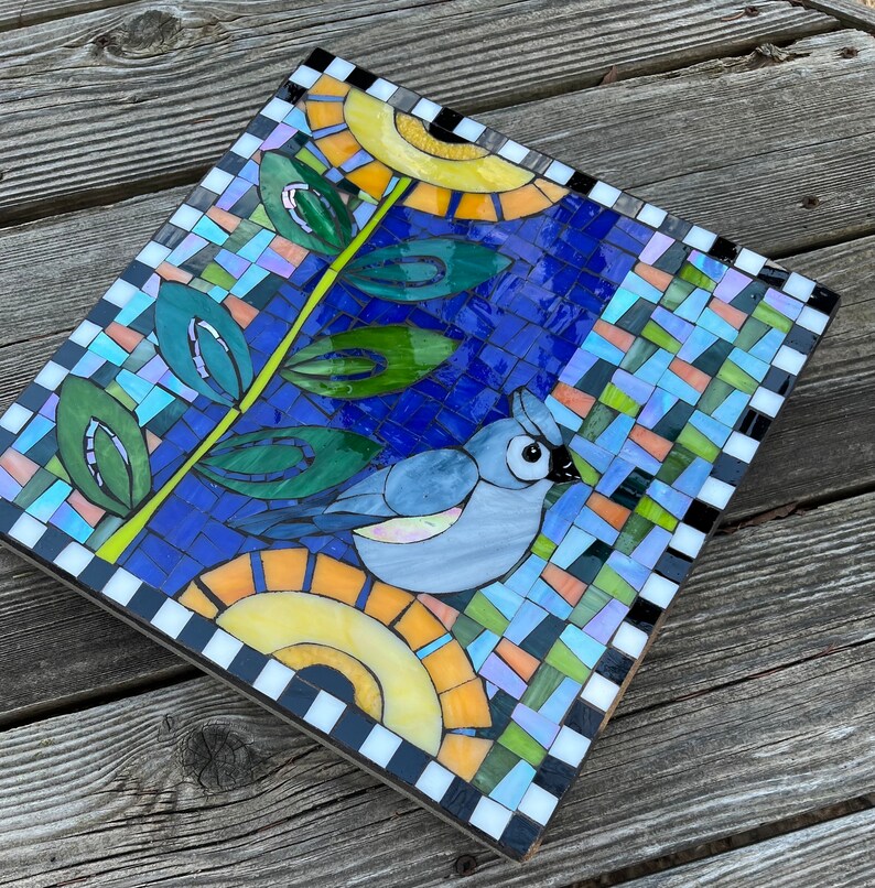 May include: Square mosaic art piece featuring a blue bird, leaves, and sun-like shapes. The mosaic is made of small, colorful tiles, with a blue background. The border is black and white checkered. The artwork is on a wooden base.