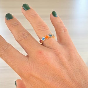 Puede incluir: Una mano con un anillo de oro con diseños de esmalte en forma de estrella de colores. La muñeca muestra varias pulseras de oro, incluida una con un patrón geométrico y detalles de piedras preciosas. Las uñas están pintadas de verde oscuro.