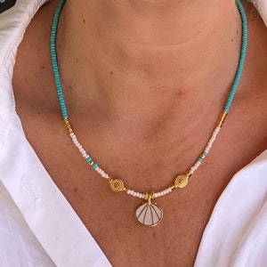 Könnte beinhalten: Eine türkisfarbene und weiße Perlenkette mit einem Muschelanhänger. Die Halskette ist mit goldfarbenen Perlen und spiralförmigen Charms verziert. Die Person trägt goldene Creolen und ein weißes Hemd.