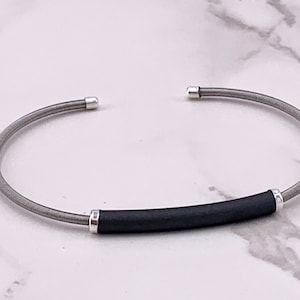 Könnte beinhalten: Ein Armreif mit einem flexiblen, gewickelten grauen Metalldrahtband. Die Mitte des Armreifs ist mit einem glatten, mattschwarzen zylindrischen Rohr akzentuiert, das auf beiden Seiten von kleinen, polierten silberfarbenen Metallkappen gehalten wird.