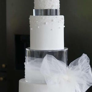 May include: A four-tier white wedding cake decorated with pearl-like sprinkles and a white tulle bow. The cake is on a clear acrylic stand.
