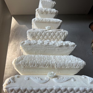 May include: A white wedding cake decorated with ruffles and flowers. The cake is shaped like a stack of pillows.