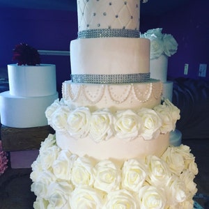May include: A white wedding cake with multiple tiers decorated with white roses, silver ribbon, and pearl accents.