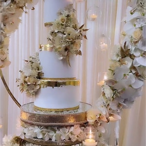 Może przedstawiać: Trzywarstwowy biały tort ślubny z złotymi akcentami i białymi kwiatami. Tort jest ozdobiony sercem z napisem "April & Trevor".