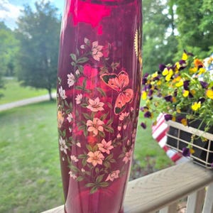 May include: A tall, ruby-colored glass vase with a gold rim. The vase is adorned with a floral and butterfly design in shades of pink, white, and green. The vase is likely used for decorative purposes.