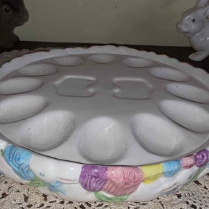 May include: A white ceramic egg holder with 12 egg-shaped indentations. The rim is decorated with pastel-colored floral and leaf designs. Two ceramic rabbit figurines are in the background.
