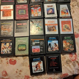 May include: A collection of twenty-two rectangular Atari video game cartridges arranged in a grid. Each black plastic cartridge features a unique colorful label with game titles such as Pac-Man, Pitfall, Defender, and Combat, showcasing various graphic illustrations.