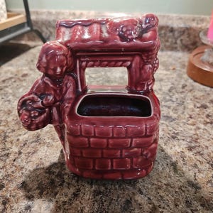 May include: A burgundy ceramic well-shaped planter. The planter features a small figure holding a basket, and is decorated with brickwork and grape clusters. The well has an opening for planting flowers or displaying small items.