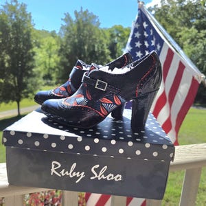 May include: A pair of black patent leather heeled shoes with a floral pattern in red and blue, sitting on a polka-dot box. The shoes have a buckle closure and a chunky heel. The box reads "Ruby Shoo".