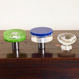 Op de afbeelding: Drie decoratieve kastknoppen. Eén is groen, één is blauw en de derde is helder. Ze hebben allemaal een zilverkleurige metalen basis en worden gepresenteerd op een donker houten oppervlak.