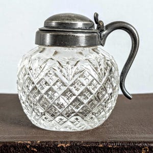 May include: A clear, round glass jar with a diamond-cut pattern. It features a silver-colored lid and handle. The jar is sitting on a brown surface. The jar is likely used for storing liquids or small items.