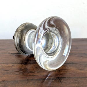 May include: A clear glass door knob featuring a smooth, rounded face and a concave center. The knob is attached to a circular, weathered metallic base with a dark, tarnished finish that connects to a cylindrical neck leading to the glass handle.