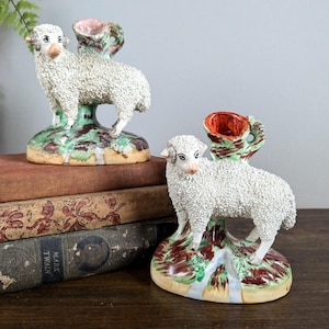 May include: Two ceramic sheep figurines with textured white wool, standing on a base with green, brown, and blue accents. Each figurine has a small, open cup on its back, with a pink interior and green exterior.