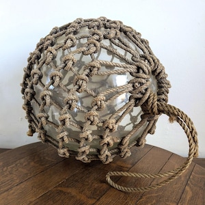 Pode incluir: Uma boia de vidro transparente, envolta em uma rede de corda bege com nós, com uma alça de corda. A boia está sobre uma superfície de madeira. Item de decoração náutica redondo e vintage.