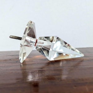 Puede incluir: Un pomo de cristal transparente y facetado con un extremo puntiagudo y una base triangular plana. El pomo tiene un eje de metal plateado con un extremo roscado para la fijación. El pomo está sobre una superficie de madera oscura.