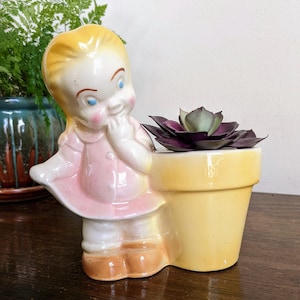 Pode incluir: Um vaso de cerâmica vintage em forma de menina, com cabelo loiro, olhos azuis e um vestido rosa. O vaso amarelo contém uma suculenta roxa. A estatueta está sobre uma superfície de madeira.