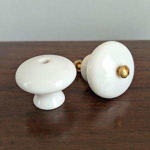 May include: Two white ceramic cabinet knobs with a glossy finish. One knob is mushroom-shaped, and the other is oval, both with gold-colored metal accents. The knobs are displayed on a dark brown wooden surface.