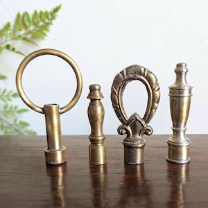 May include: Four antique brass lamp finials are arranged on a wooden surface. The finials showcase diverse designs, including a ring, a small knob, an oval with a fleur-de-lis, and a classic urn shape. The brass has a warm, aged patina.