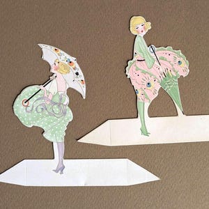 Könnte beinhalten: Zwei Vintage-Papierausschnitte von Frauen in Kleidern im Flapper-Stil. Eine Figur hält einen Sonnenschirm mit floralen Akzenten, während die andere ein rosa-grünes Kleid mit Pfauenfedermuster trägt. Beide Figuren stehen auf weißen Papierständern.