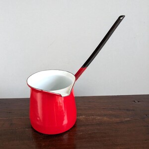 May include: A small, red enamel coffee pot with a long, black handle. The interior is white, and the rim is outlined in black. The pot sits on a dark wooden surface, with a plain, light-colored background.