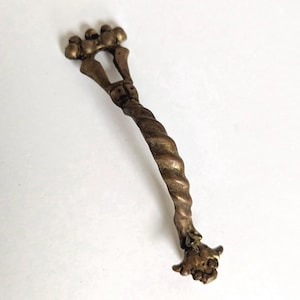 May include: A metallic, bronze-toned handle featuring a twisted, spiral-shaped central bar. The top end displays a crown-like design with four small spheres, while the bottom end terminates in a detailed, sculpted floral or animal-like ornament.